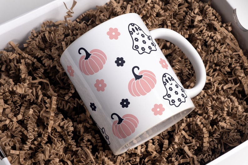 Puede incluir: Taza de cer&aacute;mica blanca con tem&aacute;tica de Halloween, con calabazas rosas, flores negras e ilustraciones de fantasmas. La taza se presenta en una caja de regalo blanca con relleno de papel marr&oacute;n.