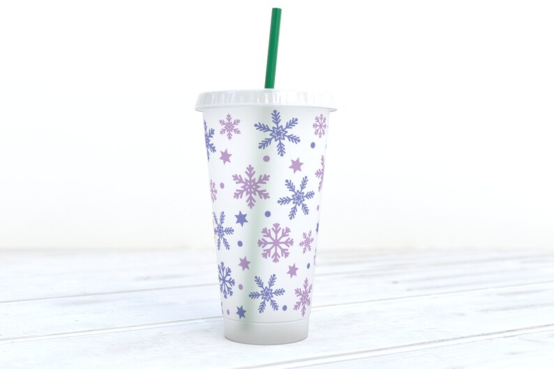 May include: A white reusable cup with a green straw. The cup has a snowflake pattern in shades of purple and blue.