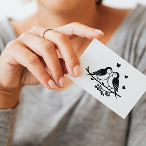 May include: A white card with a black silhouette of two birds perched on a branch with a small cluster of berries. Three small butterflies are also depicted on the card.