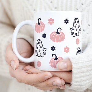 Puede incluir: Taza de cer&aacute;mica blanca con un patr&oacute;n repetido de calabazas rosas, flores negras e ilustraciones de fantasmas. La taza tiene un asa blanca y la sostiene una persona que lleva un su&eacute;ter blanco. El dise&ntilde;o es adecuado para Halloween.