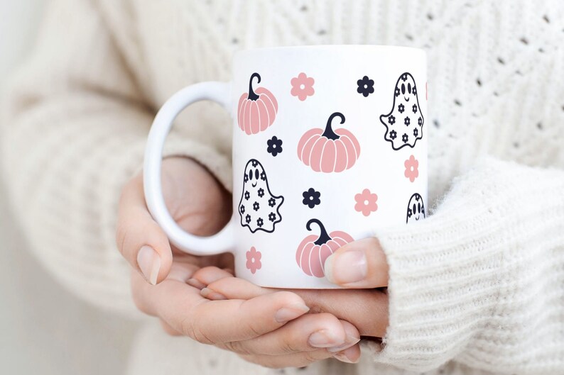 Puede incluir: Taza de cer&aacute;mica blanca con un patr&oacute;n repetido de calabazas rosas, flores negras e ilustraciones de fantasmas. La taza tiene un asa blanca y la sostiene una persona que lleva un su&eacute;ter blanco. El dise&ntilde;o es adecuado para Halloween.