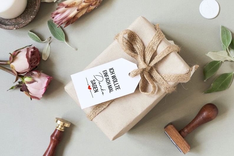 K&ouml;nnte beinhalten: Eine braune Geschenkbox, die in braunes Papier eingewickelt ist, mit einer Schleife aus Juteschnur und einem wei&szlig;en Geschenkanh&auml;nger. Der Geschenkanh&auml;nger tr&auml;gt den Text "Ich wollte einfach mal Danke sagen" mit einem roten Herzen.