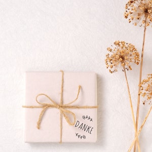 K&ouml;nnte beinhalten: Eine wei&szlig;e Geschenkbox mit einer braunen Kordelschleife und einem "Danke"-Stempel. Die Box befindet sich auf einem wei&szlig;en strukturierten Hintergrund mit getrockneten Blumen im Hintergrund.