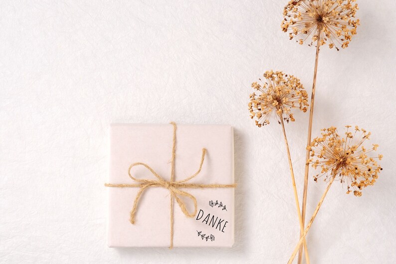 K&ouml;nnte beinhalten: Eine wei&szlig;e Geschenkbox mit einer braunen Kordelschleife und einem "Danke"-Stempel. Die Box befindet sich auf einem wei&szlig;en strukturierten Hintergrund mit getrockneten Blumen im Hintergrund.