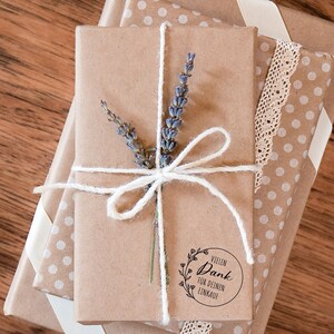 K&ouml;nnte beinhalten: Ein Stapel Geschenke, verpackt in braunes Papier mit wei&szlig;en Punkten und einem wei&szlig;en Band. Das oberste Geschenk ist mit Schnur gebunden und mit einem Lavendelzweig verziert. Ein runder Aufkleber mit dem Text "Vielen Dank f&uuml;r deinen Einkauf" befindet sich auf der Vorderseite des obersten Geschenks.