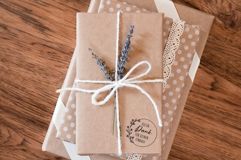 K&ouml;nnte beinhalten: Ein Stapel Geschenke, verpackt in braunes Papier mit wei&szlig;en Punkten und einem wei&szlig;en Band. Das oberste Geschenk ist mit Schnur gebunden und mit einem Lavendelzweig verziert. Ein runder Aufkleber mit dem Text "Vielen Dank f&uuml;r deinen Einkauf" befindet sich auf der Vorderseite des obersten Geschenks.