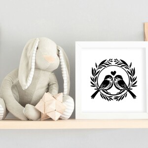 May include: A black and white silhouette of two birds in a wreath with a heart in the center. The image is framed in a white frame and displayed on a wooden shelf.