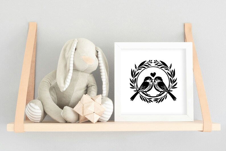 May include: A black and white silhouette of two birds in a wreath with a heart in the center. The image is framed in a white frame and displayed on a wooden shelf.
