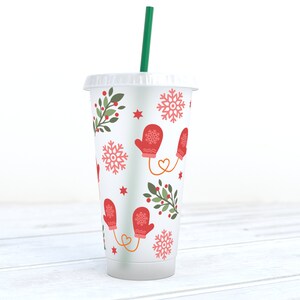 May include: A white reusable cup with a winter-themed design. The cup features red mittens, snowflakes, green holly sprigs, and red berries. A green straw is inserted in the cup.