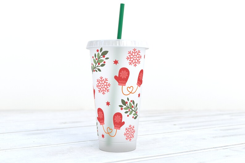 May include: A white reusable cup with a winter-themed design. The cup features red mittens, snowflakes, green holly sprigs, and red berries. A green straw is inserted in the cup.