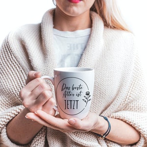 May include: White ceramic mug with a black circle and the text "Das beste Alter ist JETZT" and a small flower design.