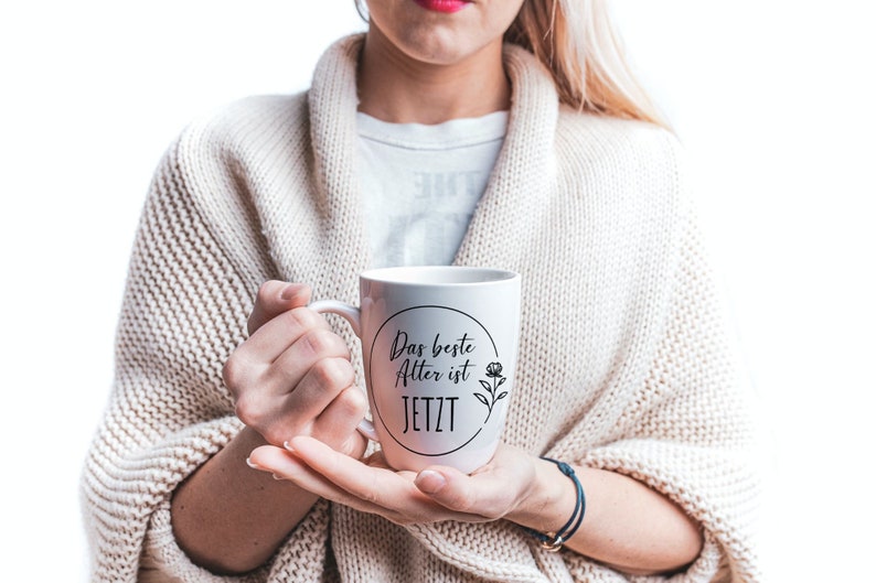 May include: White ceramic mug with a black circle and the text "Das beste Alter ist JETZT" and a small flower design.