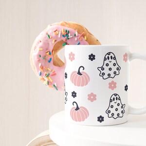Puede incluir: Una taza de cer&aacute;mica blanca con un dise&ntilde;o de Halloween con calabazas rosas, fantasmas y flores. Una rosquilla rosa con chispas de colores est&aacute; al lado de la taza. La taza tiene un asa curvada.