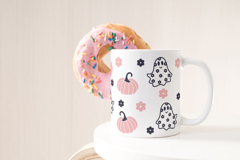Puede incluir: Una taza de cer&aacute;mica blanca con un dise&ntilde;o de Halloween con calabazas rosas, fantasmas y flores. Una rosquilla rosa con chispas de colores est&aacute; al lado de la taza. La taza tiene un asa curvada.