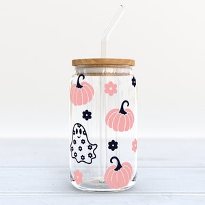May include: A clear glass jar with a wooden lid and a straw. The jar is decorated with pink pumpkins, black flowers, and a black ghost with a white polka dot pattern.