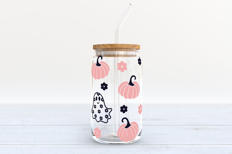 May include: A clear glass jar with a wooden lid and a straw. The jar is decorated with pink pumpkins, black flowers, and a black ghost with a white polka dot pattern.