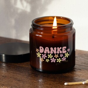 K&ouml;nnte beinhalten: Ein braunes Glas-Kerzengef&auml;&szlig; mit einem rosa-gelben Blumendesign und dem Wort "DANKE" in rosa Buchstaben. Die Kerze ist angez&uuml;ndet und das Gef&auml;&szlig; hat einen schwarzen Deckel.