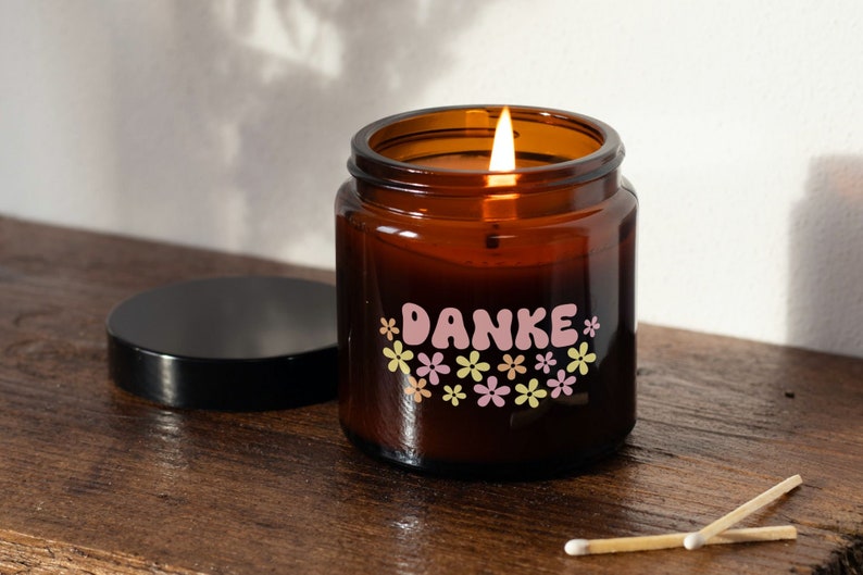 K&ouml;nnte beinhalten: Ein braunes Glas-Kerzengef&auml;&szlig; mit einem rosa-gelben Blumendesign und dem Wort "DANKE" in rosa Buchstaben. Die Kerze ist angez&uuml;ndet und das Gef&auml;&szlig; hat einen schwarzen Deckel.