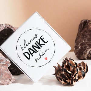 K&ouml;nnte beinhalten: Wei&szlig;e Geschenkbox mit einem schwarzen und wei&szlig;en Kreisdesign, auf dem "kleines DANKE sch&ouml;n" mit einem kleinen roten Herzen steht. Die Box steht auf einer wei&szlig;en Oberfl&auml;che mit braunen Steinen und einer getrockneten Blume.