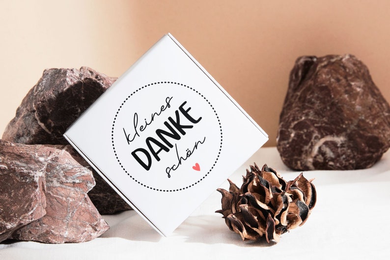 K&ouml;nnte beinhalten: Wei&szlig;e Geschenkbox mit einem schwarzen und wei&szlig;en Kreisdesign, auf dem "kleines DANKE sch&ouml;n" mit einem kleinen roten Herzen steht. Die Box steht auf einer wei&szlig;en Oberfl&auml;che mit braunen Steinen und einer getrockneten Blume.