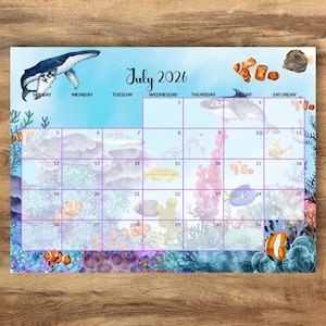 以下が含まれることがあります： 2026年7月のカレンダーページ。水彩画風の海をテーマにしており、シロナガスクジラ、クマノミ、様々なサンゴ礁の要素が描かれています。グリッドには曜日と1日から31日までの日付が表示され、水生生物が描かれた明るい青色の背景が特徴です。