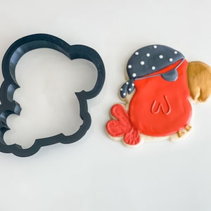 Puede incluir: Un cortador de galletas de plástico negro con forma de loro con una bandana. Junto al cortador hay una galleta decorada en rojo, dorado y blanco.