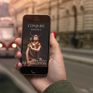 May include: A smartphone screen shows the book cover for "CONJURE Volume 1" by Rena Aliston. The cover art depicts a woman with antlers and an owl. A blurred street scene with a tram is in the background.
