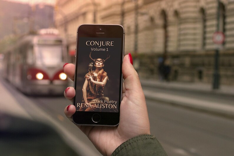 May include: A smartphone screen shows the book cover for "CONJURE Volume 1" by Rena Aliston. The cover art depicts a woman with antlers and an owl. A blurred street scene with a tram is in the background.
