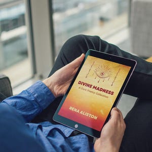 May include: A person holding a tablet displaying the book cover of "Divine Madness: A Dark Poetry Collection" by Rena Aliston. The cover features an eye illustration and text on a yellow and red background. The person is wearing a blue shirt.