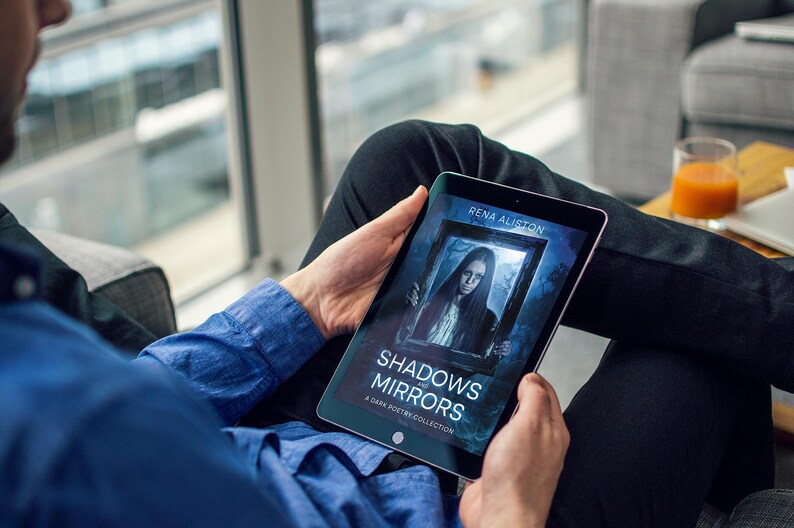 Op de afbeelding: Een tablet toont de boekomslag van "Shadows Mirrors: A Dark Poetry Collection" van Rena Aliston. De omslag toont een portret van een vrouw in een donker kader. De persoon draagt een blauw overhemd en een zwarte broek.