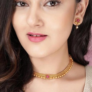 Könnte beinhalten: Eine goldene Choker-Kette mit einem rosafarbenen Stein in der Mitte. Die Kette besteht aus kleinen, runden Perlen und hat ein zartes Design. Das Model trägt dazu passende Ohrringe mit einem rosafarbenen Stein und einem Perlen-Tropfen.