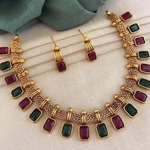 Può includere: Una collana dorata e un set di orecchini abbinati. La collana presenta un design a catena intrecciata con pietre preziose rettangolari verdi e bordeaux alternate. Gli orecchini hanno un design simile con una singola pietra preziosa bordeaux. I gioielli sono esposti su tessuto color crema.
