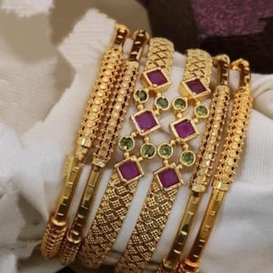 May include: A set of six gold bangles with intricate designs and pink and green gemstones. The bangles are stacked on top of each other.