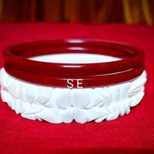 May include: Two red bangles stacked on top of a white carved bangle. The bangles are made of a smooth, shiny material.
