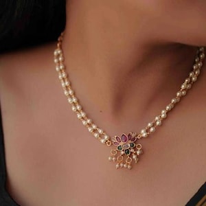 Könnte beinhalten: Eine goldene Halskette mit einer Perlenkette und einem floralen Anhänger mit bunten Edelsteinen. Der Anhänger hat ein Design aus rosa, grünen und weißen Steinen.