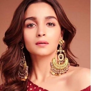 May include: A woman with long, dark brown hair wearing large, ornate gold earrings with dangling beads and coins. She is wearing a red and white polka dot top.