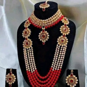 May include: A traditional Indian bridal necklace set with red and white beads, gold accents, and red stones. The set includes a long necklace, a shorter choker necklace, and matching earrings.