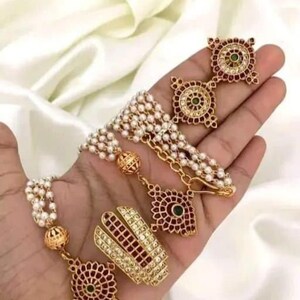 May include: A gold necklace with a pearl strand and three pendants. The pendants are a hand symbol, a round design with red and green stones, and a smaller round design with red and green stones.