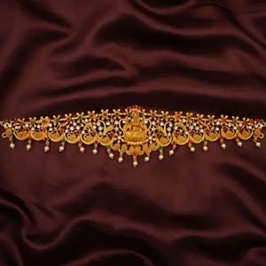 Op de afbeelding: Een gouden taillegordel met een godinfiguur in het midden, omringd door bloemen- en pauwenmotieven. De gordel is versierd met witte parels en gekleurde edelstenen.