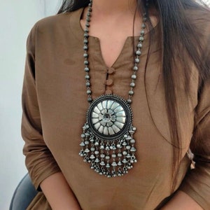 May include: A silver necklace with a large round pendant featuring a floral design and a fringe of small bells.