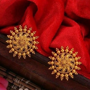 Puede incluir: Dos pendientes dorados con diseño de sol. Cada pendiente presenta un motivo floral central rodeado de pequeñas cuentas redondas. Los pendientes están sobre una tela roja vibrante.