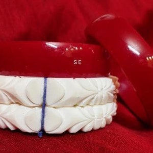 Może przedstawiać: Trzy czerwone plastikowe bransoletki ułożone na dwóch białych, rzeźbionych bransoletkach. Białe bransoletki mają kwiatowy wzór.