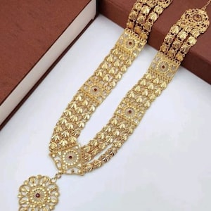 May include: A gold-coloured necklace with detailed filigree and floral patterns. The necklace has multiple strands and a large, decorative pendant with a central red stone. The jewellery is presented on a white surface with a brown box in the background.