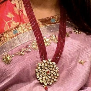 May include: A long, layered necklace with a large, round pendant. The pendant is made of gold and has a floral design with many small stones. The necklace is made of red beads and is worn by a person with a pink and red sari.