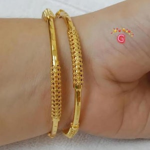 May include: Two gold bangles with intricate designs. The bangles are stacked on top of each other and have a shiny finish.