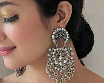 May include: Silver tone dangle earrings with a large, ornate, geometric design featuring a central, clear, faceted stone. The earrings have a crescent moon shape at the top and a fringe of small beads.