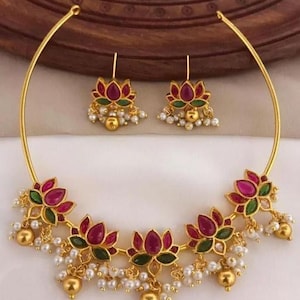May include: A gold-coloured necklace and earring set. The necklace and earrings feature lotus flower designs with red and green petals, accented with small white pearls and gold beads. The jewellery is displayed on a white surface.