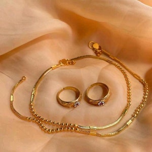 May include: A gold jewellery set comprising two rings with small pink gemstones and a delicate necklace. The set is displayed on peach-coloured fabric, highlighting the intricate details. The rings and necklace are made of gold.