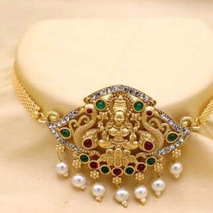 Op de afbeelding: Een gouden chokerketting met een hanger met een godheidfiguur omringd door pauwen en groene en rode edelstenen. De hanger is versierd met witte parels.