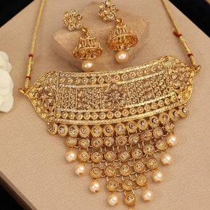 Könnte beinhalten: Ein goldfarbenes Halsketten-Set mit einem großen, verzierten Choker und baumelnden Perlenakzenten. Das Set enthält auch passende Ohrringe mit Perlenanhängern.